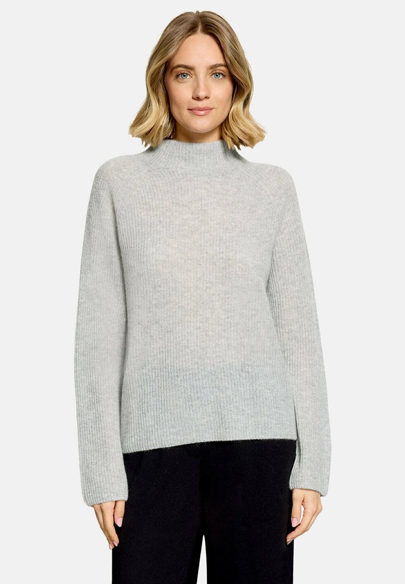 Femme aux cheveux blonds mi-longs portant un pull en maille gris clair à col montant et un pantalon noir, debout devant un fond uni.