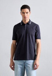 Marineblå polo med to-knaps placket og hvit stripet krage. Mykt bomullsmateriale. Brodert logo på brystet, sammen med lyse denimjeans.