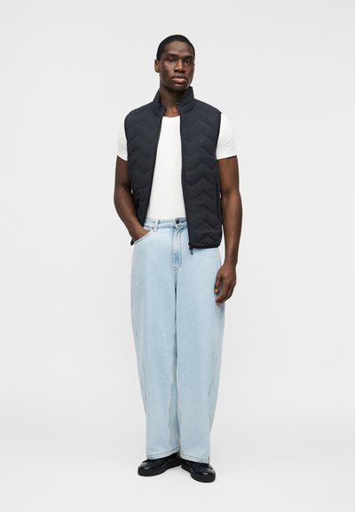 Άνδρας που στέκεται, φοράει μαύρο γιλέκο με ραφές πάνω από ένα λευκό μπλουζάκι, ανοιχτόχρωμο μπλε τζιν με φαρδιές παντελόνες και μαύρα παπούτσια, με τα χέρια στις τσέπες.