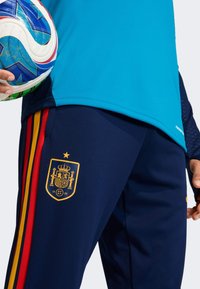 Blå atletisk skjorte kombinert med marineblå bukser med røde og gule striper, gyltne Spania-emblem og teksturert stoff. Fotball holdt i hånden.