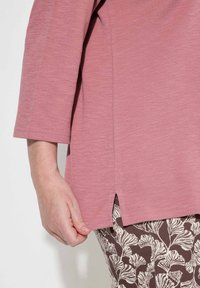 Rosa Langarmoberteil mit strukturiertem Finish, ausgestattet mit einem Seitenschlitz, kombiniert mit braunen Leggings mit Muster, die blätterartige weiße Designs zeigen.