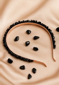 Czarna opaska na włosy z cienką metalową podstawą, ozdobiona wypolerowanymi czarnymi kamieniami na górze. Kilka luźnych czarnych kamieni na beżowym tle z tkaniny.