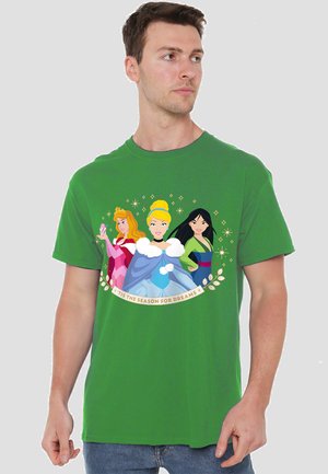 Junger Mann trägt ein grünes T-Shirt mit drei Disney-Prinzessinnen und dem Text "Es ist die Zeit der Träume" darunter.