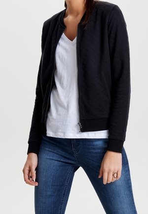 Femme portant une veste matelassée noire zippée sur un t-shirt blanc à col en V et un jean bleu, debout devant un fond uni.