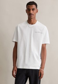 Witte katoenen t-shirt met een ronde hals, korte mouwen en een subtiele groene logo op de borst. Draagt met zwarte jeans.