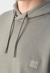 Gros plan d'un sweat à capuche gris-vert avec des cordons et un petit patch brodé du logo BOSS sur la poitrine.