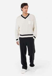 White ribbed sweater with black V-neck and cuffs, featuring a round emblem. Worn with black trousers and white sneakers.
