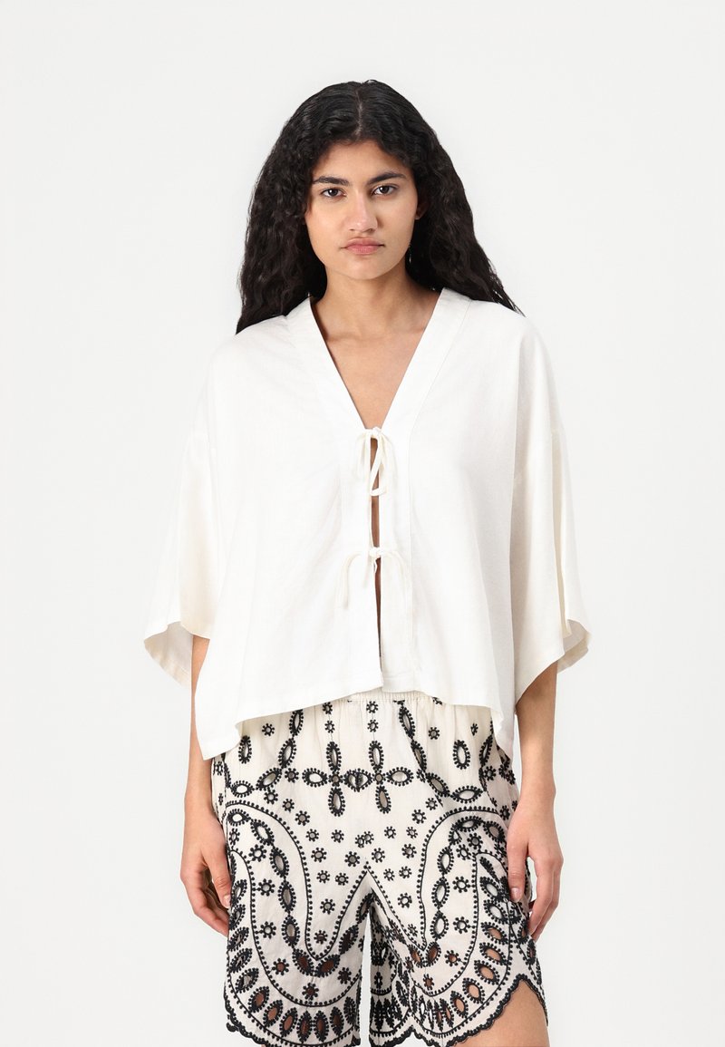Hvit cropped topp med dyp V-hals og snor-detalj; kombinert med mønstrete svart-hvite shorts med blomsterbroderi.