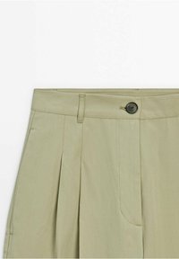 Pantalon beige clair taille haute avec plis, un seul bouton noir, passants pour ceinture et une fermeture frontale à glissière dissimulée.