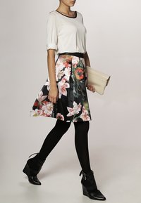 Jupe à imprimé floral noire avec des accents roses et verts, associée à un haut blanc et des collants noirs, tenant une pochette beige. Bottines à talons hauts.