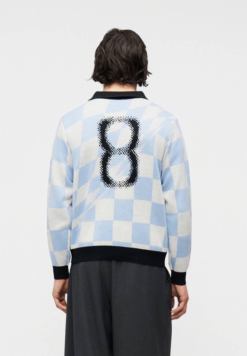 Άτομο με κοντά σκουρόχρωμα μαλλιά, που φοράει ελαφρύ γαλάζιο και λευκό πουλόβερ με καρό μοτίβο και μεγάλο ψηφιακό αριθμό 8 στην πλάτη.