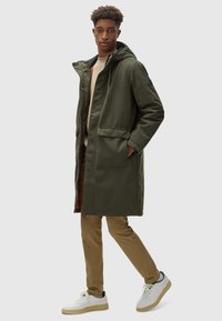 Parka con capucha en color verde oliva, con cremallera frontal, grandes bolsillos y una silueta ligeramente ajustada, llevada sobre un suéter beige y pantalones beige.