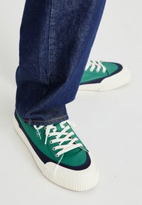 Zelené a tmavě modré plátěné tenisky s bílými gumovými podrážkami a očky, které mají bílé tkaničky a texturovaný pruhovaný vzor po stranách.
