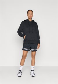 Μαύρη φούτερ με τσέπη μπροστά και κορδόνια, συνδυασμένη με μαύρες βερμούδες με λευκές ρίγες, λευκές κάλτσες και αθλητικά παπούτσια.