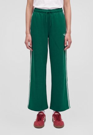 Modèle portant un pantalon de survêtement Adidas vert à jambes larges, avec des bandes blanches sur les côtés et une taille à cordon, assorti à des baskets rouges.