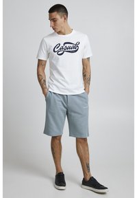 T-shirt blanche avec impression marine "Casual", associée à un short bleu clair. Le modèle porte des baskets noires. Manches courtes, coupe décontractée.