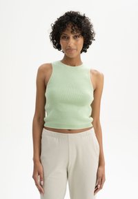 Helles grünes, geripptes Strick-Crop-Top mit hohem Kragen und ärmellosem Design, kombiniert mit beigen, lässig geschnittenen Hosen. Glatte Textur.