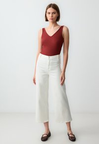 Body rosso a coste con scollatura a V, abbinato a jeans bianchi a gamba larga e scarpe basse marrone scuro, su sfondo neutro.
