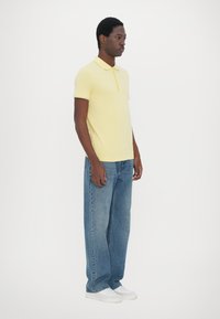 Světlě žluté polokošile s límečkem a knoflíkovou légou, v kombinaci s volnými džínovými kalhotami. Každý outfit doplňují bílé tenisky.