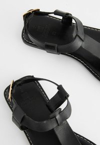 Mustat nahka sandaalit littealla pohjalla, joissa on säädettävät hihnat ja kultasävytteiset yksityiskohdat. Huomattava valkoinen tikkaus reunoilla.