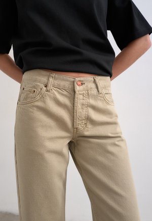 Άτομο που φοράει μπεζ τζιν με ψηλή μέση, κουμπί από χαλκό και τσέπες, σε συνδυασμό με μια μαύρη κοντομάνικη πουκάμισο μέσα στην μπλούζα.