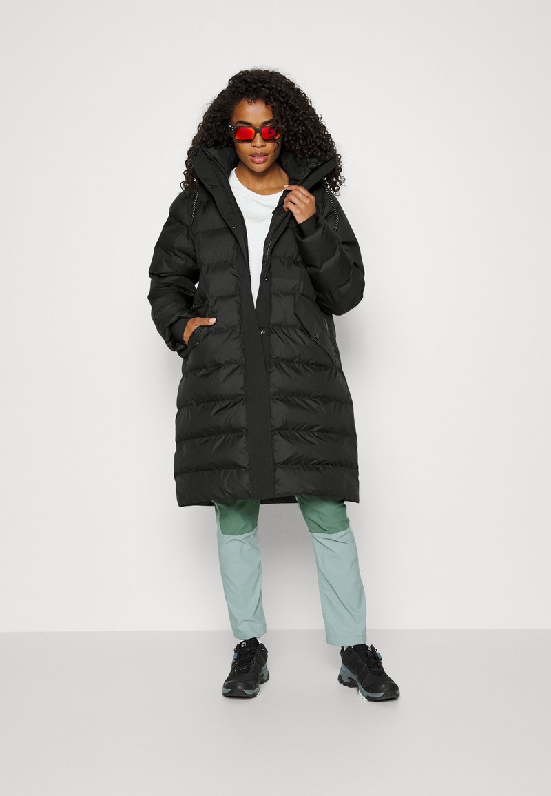 Manteau matelassé noir avec fermeture éclair sur le devant et poches latérales, doté d'un col haut. Le modèle l'a associé à un pantalon vert clair et des baskets noires.