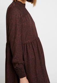 Brun mönstrad klänning med långa ärmar, hög hals med smockdetalj, och en utställd siluett med gathers vid midjan.