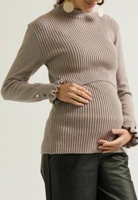 Zwangere vrouw in een geribbelde mauve trui en zwarte broek, die haar babybuik voorzichtig met beide handen vasthoudt.