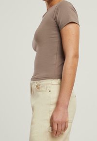 Dopasowana brązowa koszulka na krótki rękaw z miękkiego materiału, w parze z jasnobeżowymi spodniami, zaprezentowana z boku, aby podkreślić kształt i dopasowanie.