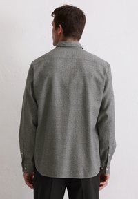 Mężczyzna w szarym, fakturowanym longsleeve z zapiętymi mankietami, widziany z tyłu na tle gładkiej białej ściany.
