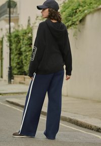 Black hooded sweatshirt with a large number 3 on the sleeve, paired with navy wide-leg trousers featuring white side stripes. Casual footwear.