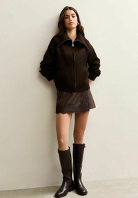 Brązowa kurtka z fakturą na zamek, dopasowana brązowa skórzana spódnica z koronkowym wykończeniem oraz brązowe botki do kolan, modelka stojąca na tle beżowej ściany.