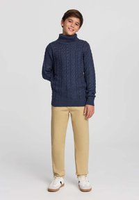 Granatowy sweter z golfem wykonany z dzianiny, zdobiony wzorem w warkocze, zestawiony z beżowymi spodniami i białymi trampkami z czarnymi akcentami.