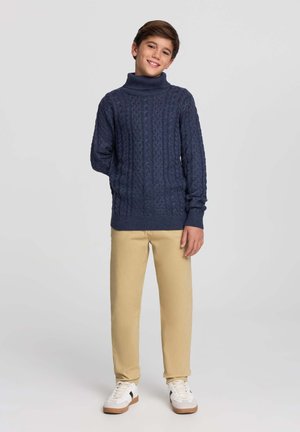 Maglione a collo alto lavorato a maglia di colore blu navy con motivo a treccia, abbinato a pantaloni beige e sneakers bianche con dettagli neri.