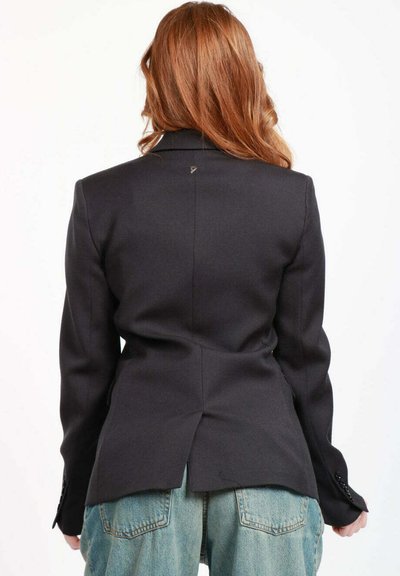 Blazer nero su misura con spalle strutturate, vita aderente, spacco posteriore e polsini abbottonati. Il materiale sembra liscio, senza schemi visibili.