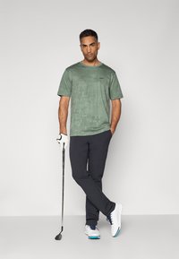 Chemise de golf à motifs verts avec des manches courtes, associée à un pantalon bleu marine. Chaussures de golf blanches avec des accents bleus et un club de golf en main.