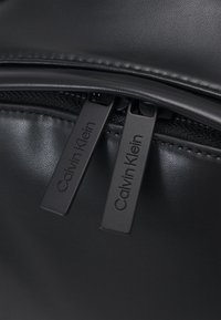 Schwarzer Rucksack aus synthetischem Leder mit zwei schwarzen Reißverschlüssen, auf denen "Calvin Klein" eingraviert ist. Glatte Textur, minimalistisches Design und keine sichtbaren Muster.