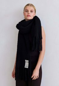 Blonde vrouw in een mouwloze zwarte jurk en een grote zwarte sjaal met franjes en twee zichtbare witte etiketten.
