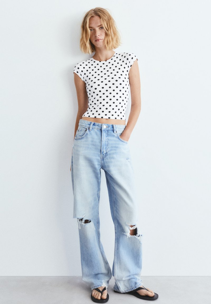 Jeune femme aux cheveux blonds ondulés porte un crop top blanc à pois noirs, un jean bleu clair déchiré et des tongs noires.