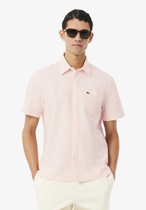 Hombre con gafas de sol oscuras, camisa de manga corta color rosa claro con bolsillo y pantalones de color crema, de pie con las manos en los bolsillos.