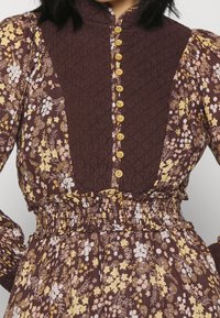 Robe florale marron avec un corsage texturé, des détails à boutons et une taille froncée. Dispose de motifs floraux jaunes et blancs sur les manches.