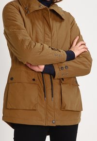 Brun isolerad jacka med dragkedja fram, knappdetaljer, stora framfickor och dragsko i midjan. Har snapkrage och quiltat mönster.