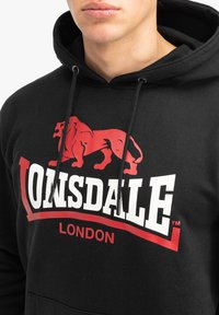 Sudadera negra con un gráfico de león rojo y el texto "Lonsdale London". Cuerdas con puntas metálicas plateadas. Textura de tela suave.