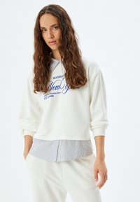 Sudadera blanca con una textura acanalada y texto azul de "New York"; presenta un cuello y dobladillo con detalles a rayas. Combinada con pantalones blancos.
