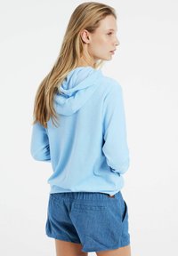 Felpa con cappuccio di colore azzurro chiaro, dal texture morbido, con maniche lunghe e vestibilità rilassata, abbinata a pantaloni corti blu.