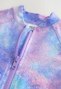 Vêtement zippé dans des couleurs pastel arborant un motif galactique avec des points métalliques, fabriqué en tissu lisse avec une texture douce.
