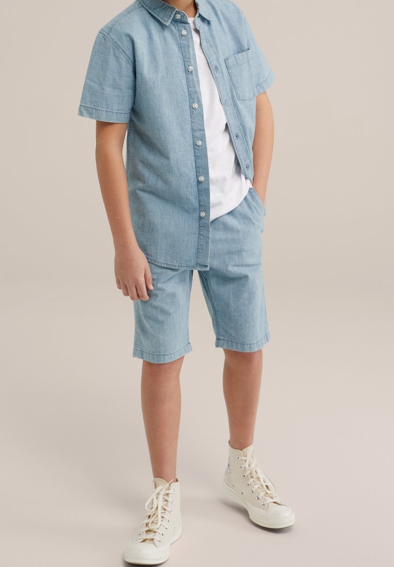 Světle modrá riflová košile s krátkým rukávem, s knoflíkovou přední částí a náprsní kapsou, spárovaná s odpovídajícími šortkami. Bílé tričko pod tím, béžové vysoké tenisky.