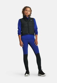 Gilet imbottito nero sopra una maglietta a maniche lunghe blu, abbinato a leggings blu e neri. Scarpe da ginnastica nere con suole texturate.