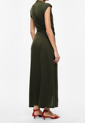 Donna in piedi con abito lungo verde oliva senza maniche e sandali rossi con tacco basso su sfondo bianco, di spalle.