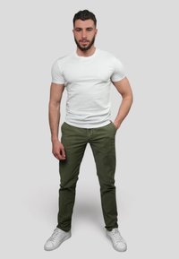 Un t-shirt blanc à manches courtes associé à un pantalon vert olive et à des baskets blanches, présenté sur un modèle masculin sur fond gris neutre.